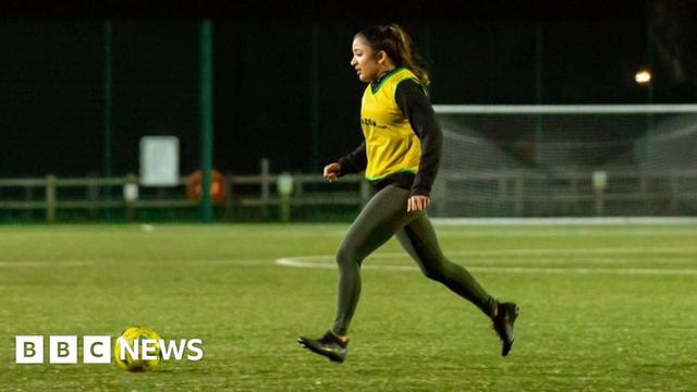 A women stands on a football pitch kicking a football. It is very dark and she is wearing a yellow T-shirt and black leggings