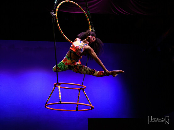 Summer Circus Celebration - aerialist Darielle performs an act from the show The RAiNBOW is Enuf. She is on an invented apparatus called "aerial drum" with a tippy lyra above and two smaller hoops below that resemble the outline of an African djembe drum. The hoops are wrapped with yellow, green and red tape. She is hanging from the upper lyra by one arm, while her other arm is holding one leg up in a splits position. She is wearing abstract patterned tights and a top in red, yellow, green, black, and blue colors.