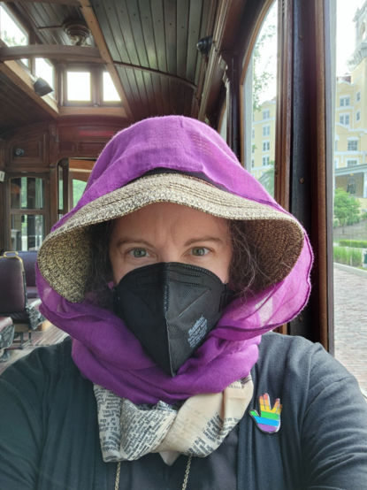 A woman dressed in a black shirt with a rainbow "Vulcan salute" pin, black N95 mask, and a wide-brimmed straw hat tied on by a purple gauze scarf takes a selfie inside an antique trolley.  Along the center of the wood-lined ceiling is a raised section with little square windows.  Out the windows over her left shoulder can be seen the yellow exterior of the West Baden Springs Hotel.