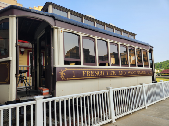 Exterior of an antique trolley with slightly arched windows along the length of it, with a raised central section with rectangular windows.  On the side is a burgundy strip with (painted in gold) 1 FRENCH LICK AND WEST BADEN 1