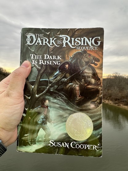 Well worn front cover of The Dark is Rising by Susan Cooper, showing a man in a dark hooded cloak riding a black horse and a boy with a strangely glowing belt buckle 