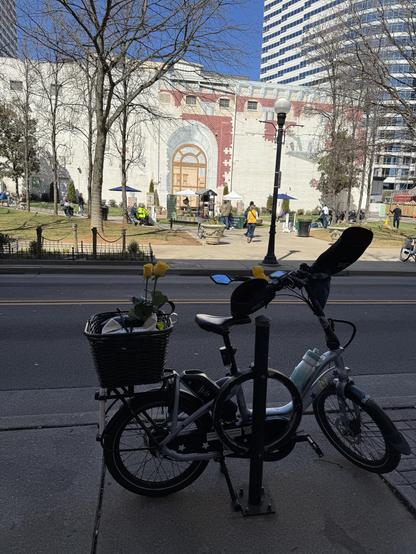 Bike parked next to a street, with a park across the way. In the distance, a singer with a guitar is doing a concert. 