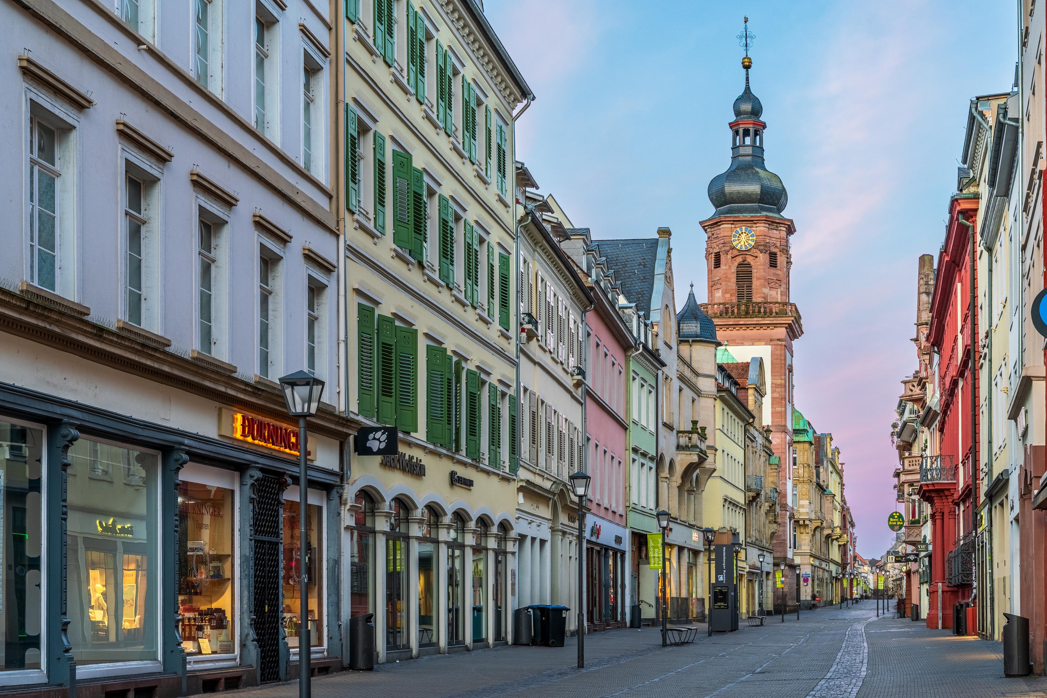 Hauptstrasse in Heidelberg. The cobbled pedestrian street is lined with Baroque pastel houses. A sandstone church a half a block away has a rounded spire that stretches into a cotton candy colored sky. Hauptstrasse in Heidelberg. The cobbled pedestrian street is lined with Baroque pastel houses. A sandstone church a half a block away has a rounded spire that stretches into a cotton candy colored sky.